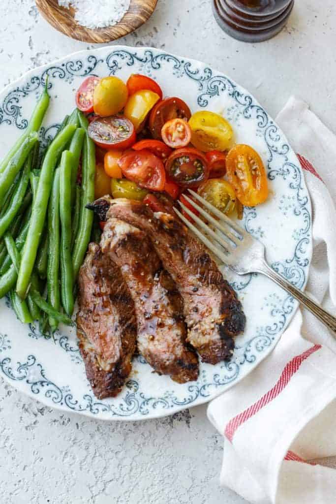 Close up of grilled Ribeye Steak served with green beans and tomatoes on a blue and white plate with a fork