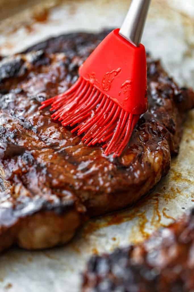 Close up of a grilled ribeye steak being rubbed with a Bar B-Q glaze with a red brush