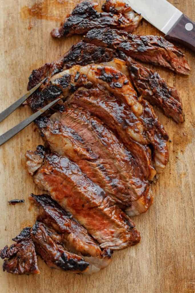 Close up of carved Grilled Ribeye Steak topped with Tequila Barbecue Glaze sitting on a wooden cutting board with a carving knife and fork