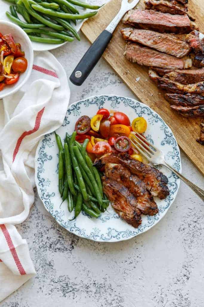 Grilled Ribeye Steak Recipe with Tequila Barbecue Glaze served with green beans and tomatoes on a blue and white plate with a fork and carved ribeye steak strips on a wooden cutting board in the background