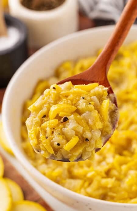 A large wooden spoon digging into sauteed yellow squash in a white bowl