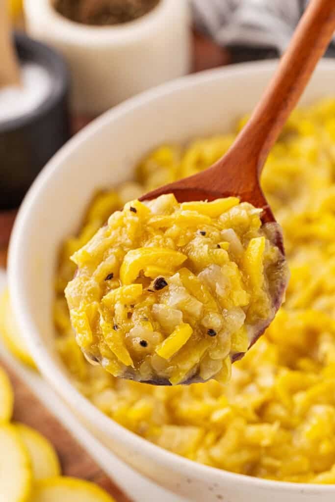A large wooden spoon digging into sauteed yellow squash in a white bowl