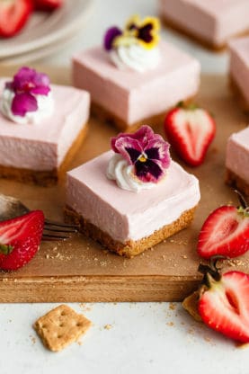 Close up of several pieces of no bake strawberry cheesecake on top of a wooden cutting board and topped with whipped cream and flowers
