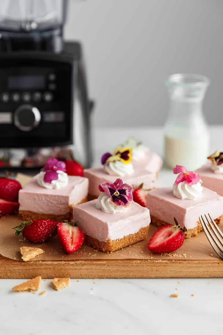 Several pieces of no bake strawberry cheesecake on top of a wooden cutting board with pieces of graham crackers, sliced strawberries and a fork and a bottle of milk and a blender in the background