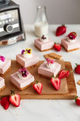 Several pieces of no bake strawberry cheesecake on top of a wooden cutting board with pieces of graham crackers, sliced strawberries and a fork and a bottle of milk and a blender in the background