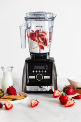 A blender filled with the ingredients to make this no bake strawberry cheesecake with graham crackers, strawberries and a glass of milk in the foreground