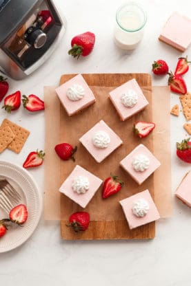Overhead shot of several pieces of no bake strawberry cheesecake on top of a wooden cutting board with pieces of graham crackers, sliced strawberries and a fork and a bottle of milk and a blender in the background