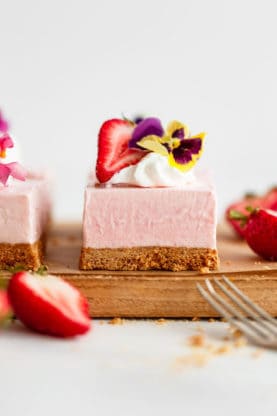 Close up of one piece of no bake strawberry cheesecake topped with whipped cream, a flower and a sliced strawberry on a wooden cutting board with a more pieces next to it