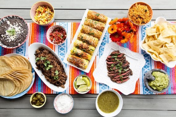Overhead shot of a picnic table full of Mexican dishes including tortillas, Mexican corn on the cob, salsa, guacamole, beans and rice