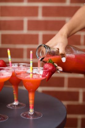 Strawberry margaritas being poured into glasses with straws on a brick background