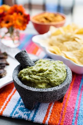 Close up of a bowl full of guacamole with a tortilla chips, rice and an arrangement of flowers in the background