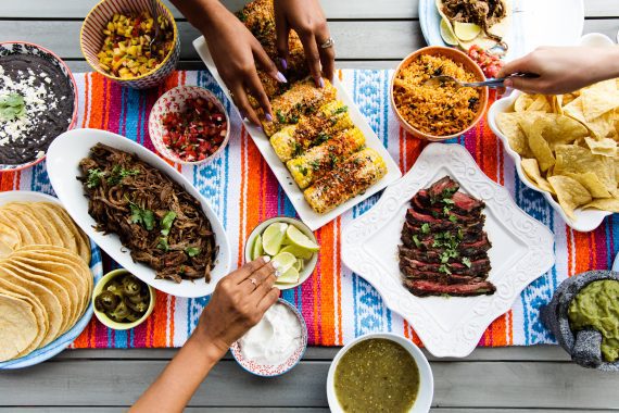 Overhead shot of a picnic table full of Mexican dishes including tortillas, Mexican corn on the cob, salsa, guacamole, beans and rice and hands grabbing food