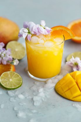 A small glass of mango citrus fruit punch garnished with flowers and an orange wedge next to mango, lime and oranges