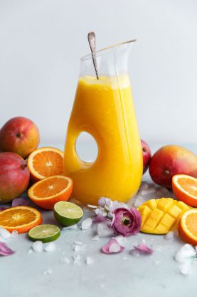 A glass pitcher full of Mango Citrus Fruit Punch Recipe with a spoon in it and surround by fresh mangoes, oranges, limes and flower petals