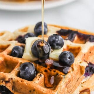 Pouring maple syrup over the top of a stack of blueberry waffles.