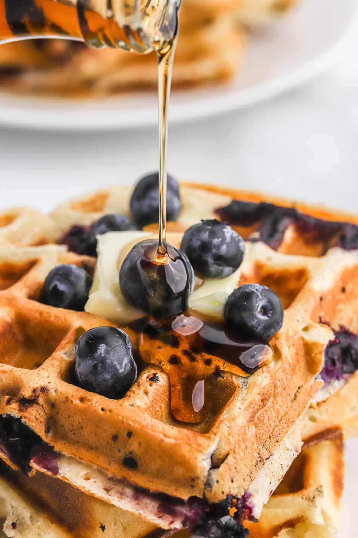 Pouring maple syrup over the top of a stack of blueberry waffles.