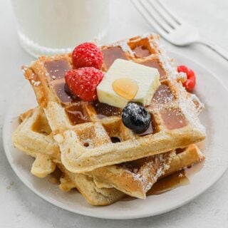 A stack of buttermilk waffles on a plate topped with a pat of butter and sliced fresh fruit.