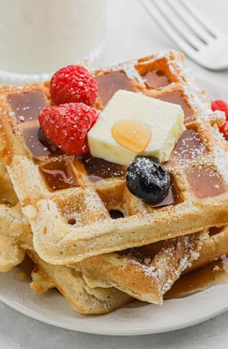 A stack of buttermilk waffles on a plate topped with a pat of butter and sliced fresh fruit.