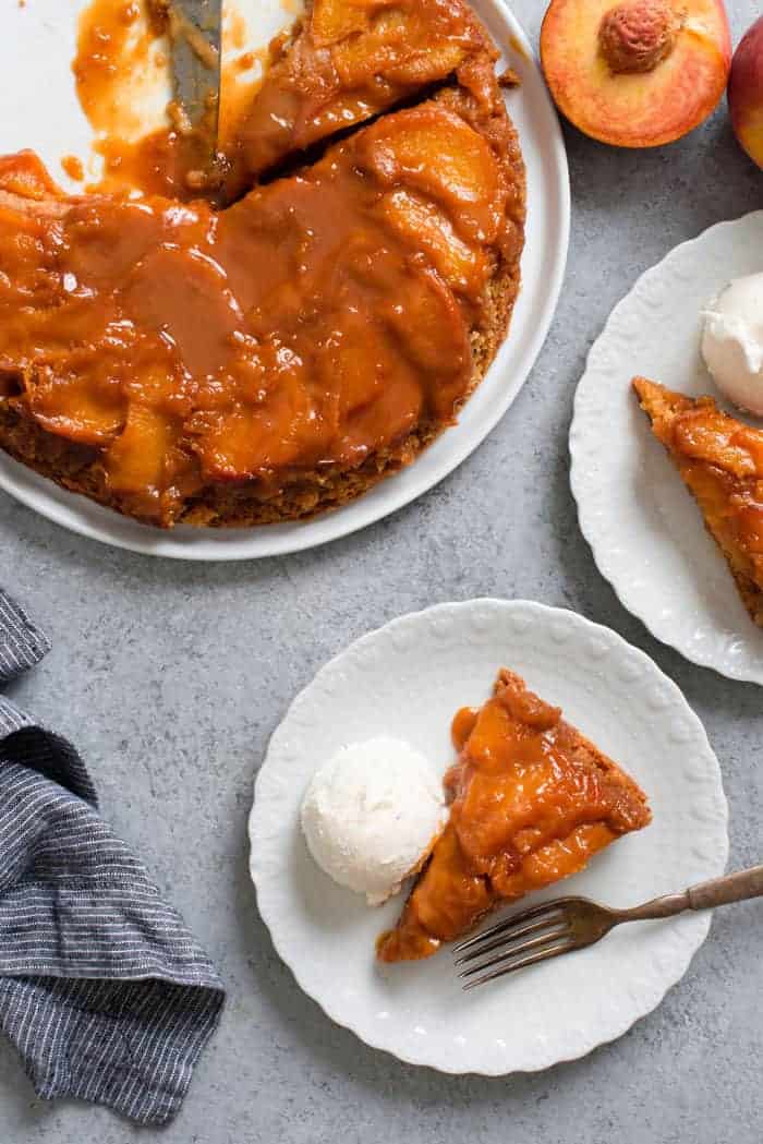 Overhead shot of caramel peach dump cake with slices missing topped with rich decadent caramel sauce and ripe tender peaches next to two slices served on white plates with a scoop of vanilla ice cream and a fork