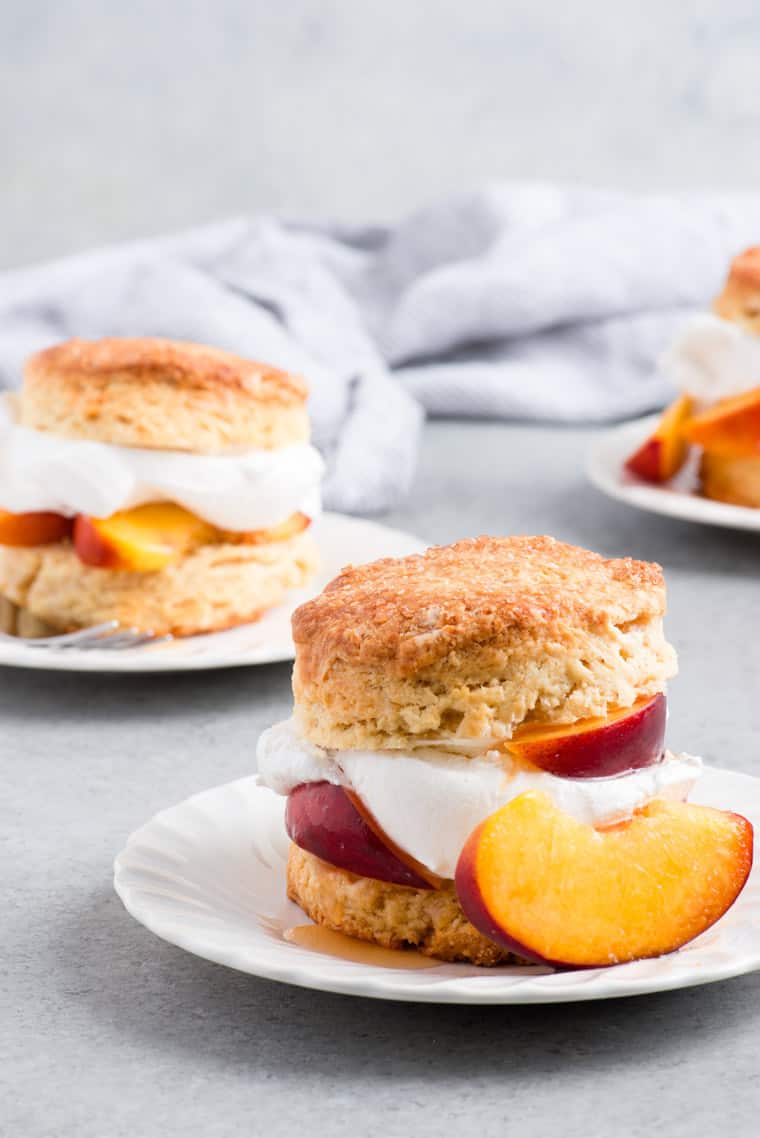 Close up of one Brown Sugar Peach Shortcake served on a white plate with a peach slice and more shortcakes in the background