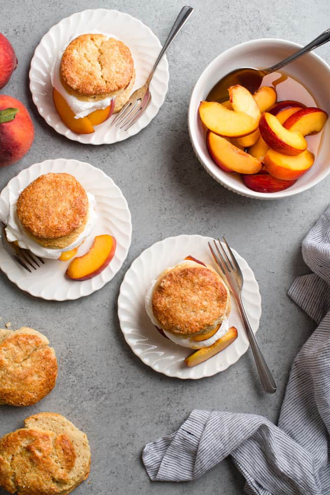 Overhead shot of three brown sugar peach shortcakes served on white plates with forks next to biscuits, fresh peaches and a bowl of sliced peaches with a spoon