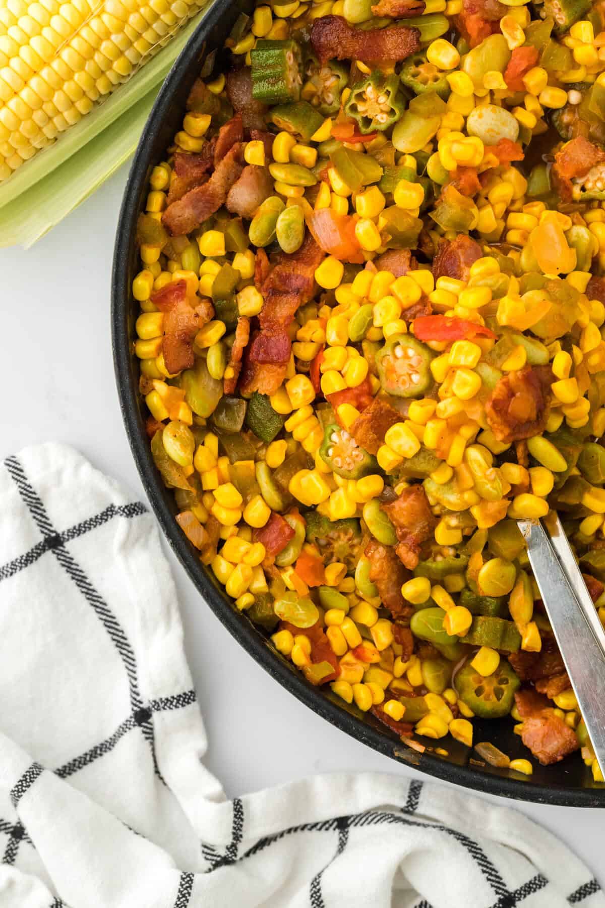 A pan filled with the best succotash recipe against a white background with a corn ear and a white napkin with black stripes