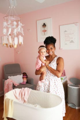 Jocelyn holding Baby Cakes Harmony in her baby nursery standing behind a Snoo machine and pink walls and paintings in the background