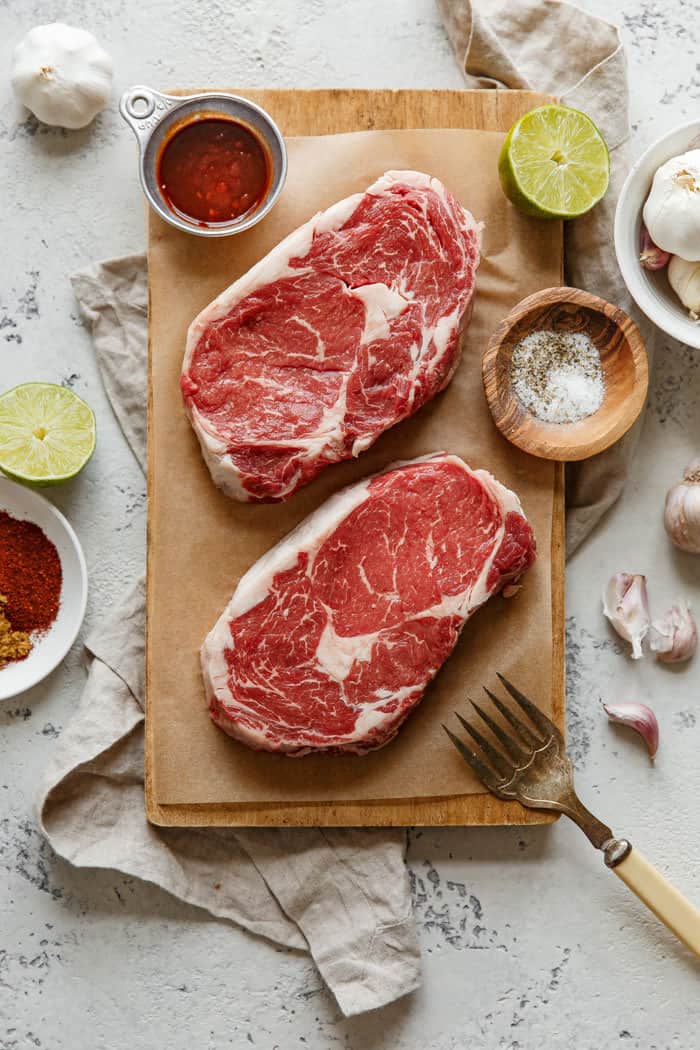 Overhead shot of Ribeye steaks ready to season and sear or grill on top of parchment paper and a wooden cutting board with steak seasoning, lime and a fork