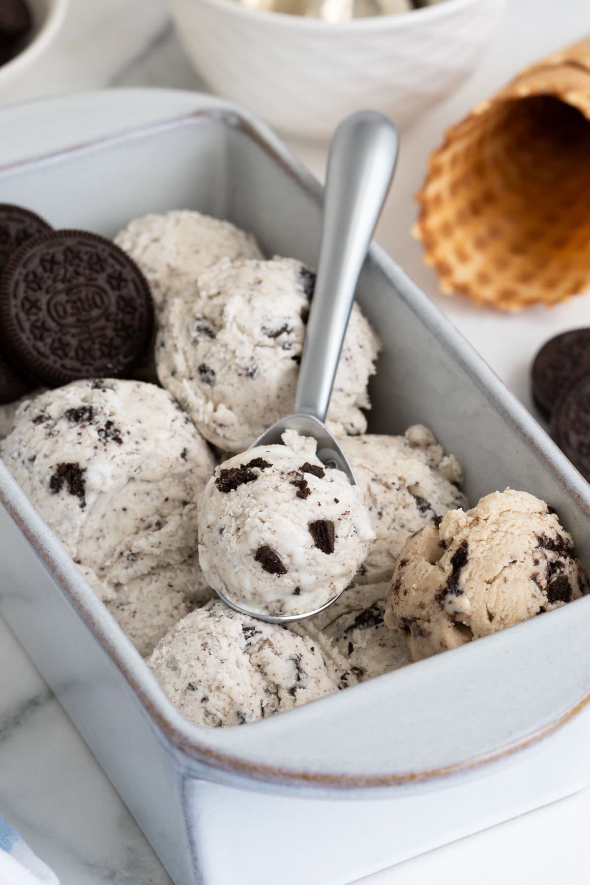 A metal ice cream scoop lifting a scoop of cookies and cream ice cream from a loaf pan, with visible cookie chunks