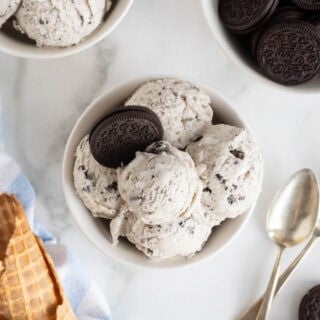 A white bowl filled with scoops of cookies and cream ice cream, garnished with a whole Oreo cookie, placed beside waffle cones and spoons