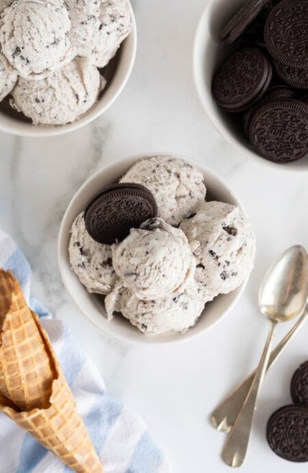 A white bowl filled with scoops of cookies and cream ice cream, garnished with a whole Oreo cookie, placed beside waffle cones and spoons