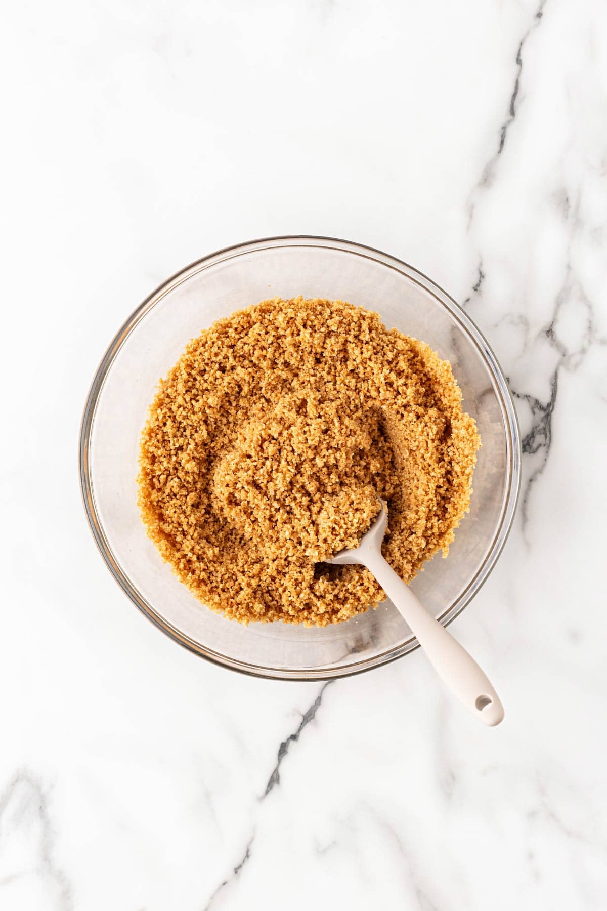 Graham cracker crust mixture in a glass bowl with a spatula on a white marble countertop