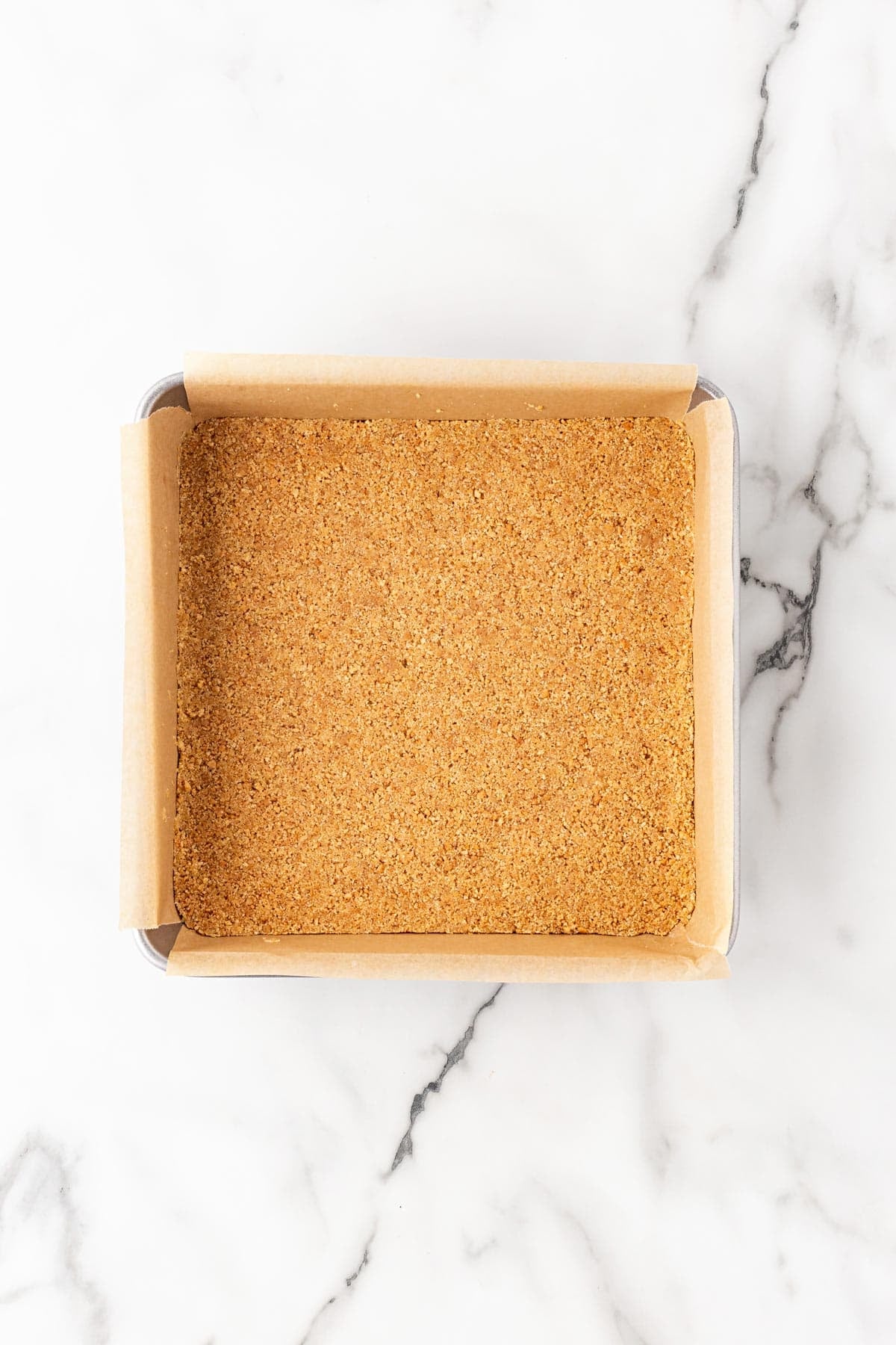 Pressed graham cracker crust in a parchment-lined baking pan on a white marble countertop