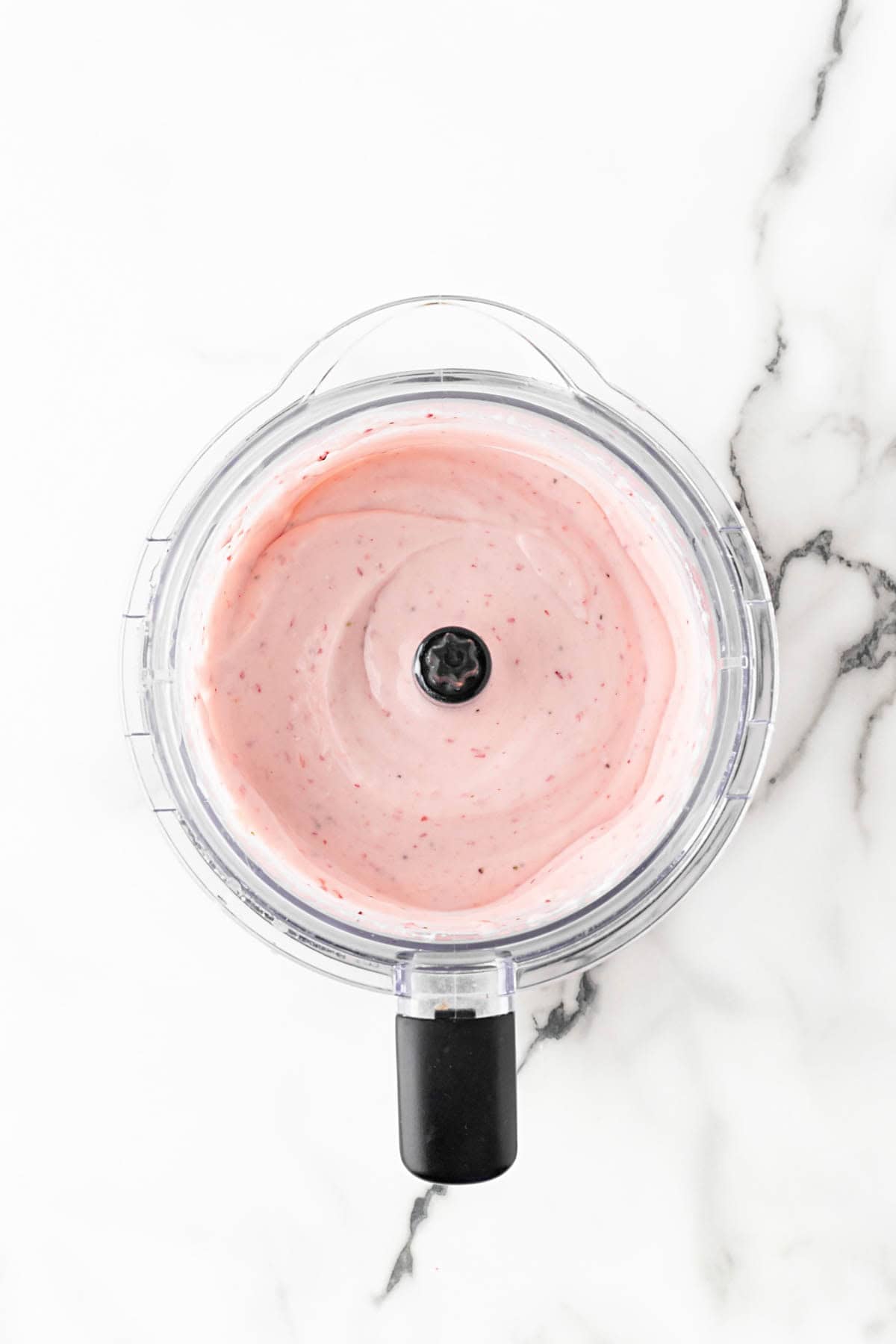 Blended strawberry cheesecake filling in a food processor on a white marble countertop