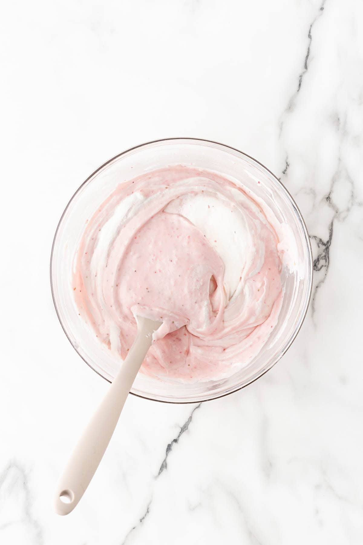 Strawberry cheesecake mixture and whipped cream folded together in a glass bowl with a spatula on a white marble countertop