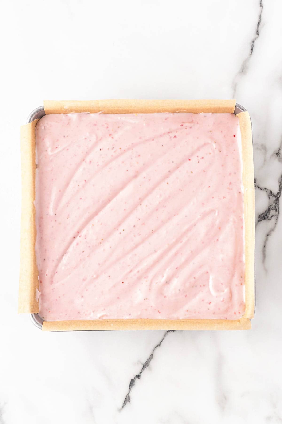 Strawberry cheesecake filling spread over a graham cracker crust in a parchment-lined pan on a white marble countertop