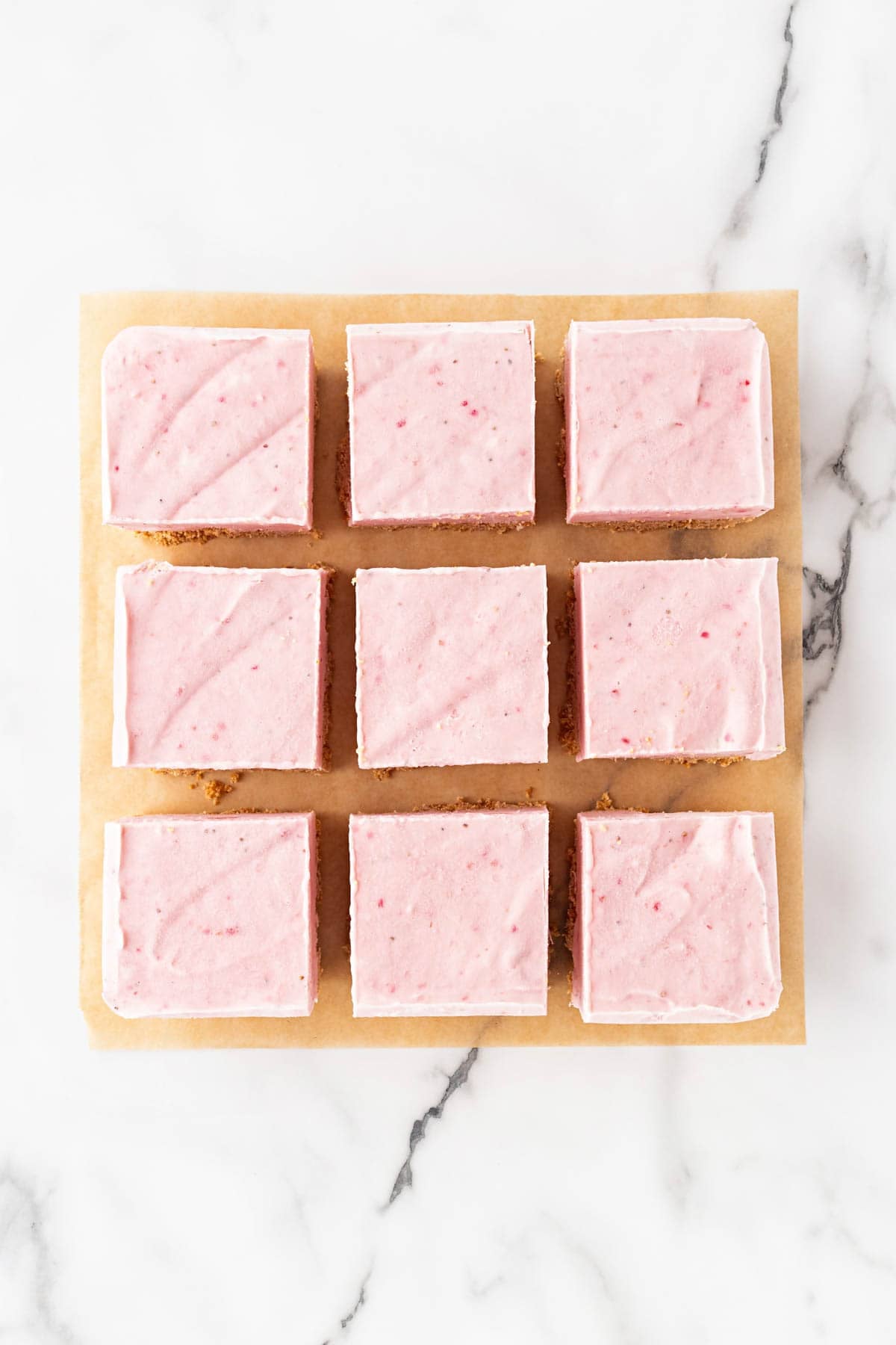 Cut strawberry cheesecake bars arranged on parchment paper on a white marble countertop