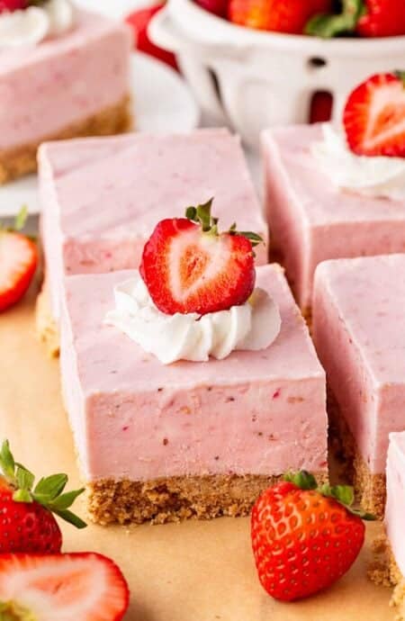 Strawberry cheesecake bars topped with whipped cream and fresh strawberries, surrounded by whole and sliced strawberries