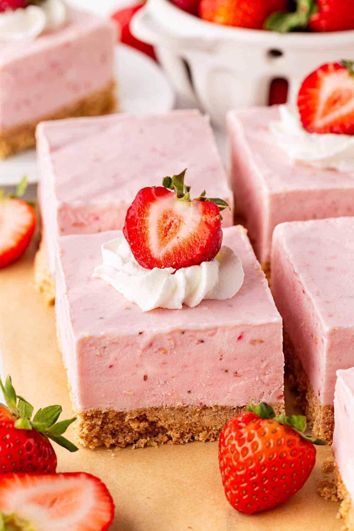Strawberry cheesecake bars topped with whipped cream and fresh strawberries, surrounded by whole and sliced strawberries