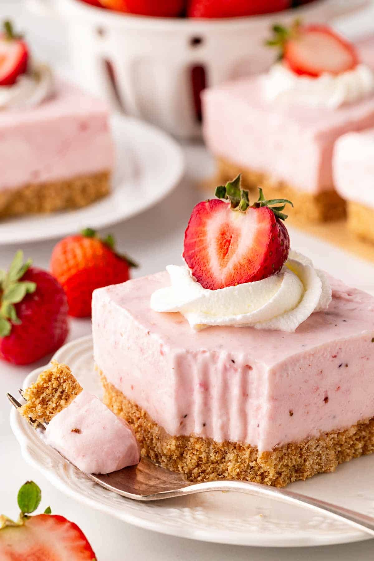 Strawberry cheesecake bar topped with whipped cream and a sliced strawberry, with a fork holding a bite and more bars in the background
