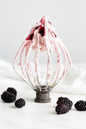 A metal whisk from a KitchenAid electric mixer with remnants of the blackberry buttercream on it and surrounded by fresh blackberries