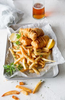 Fish on top of a bed of fried with lemons on top of parchment paper on a metal tray with a glass vinegar in the background