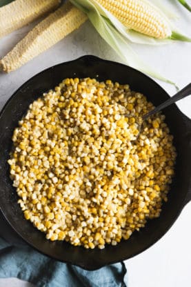 Overhead shot of fried corn in a black cast iron skillet next to shucked corn on the cob
