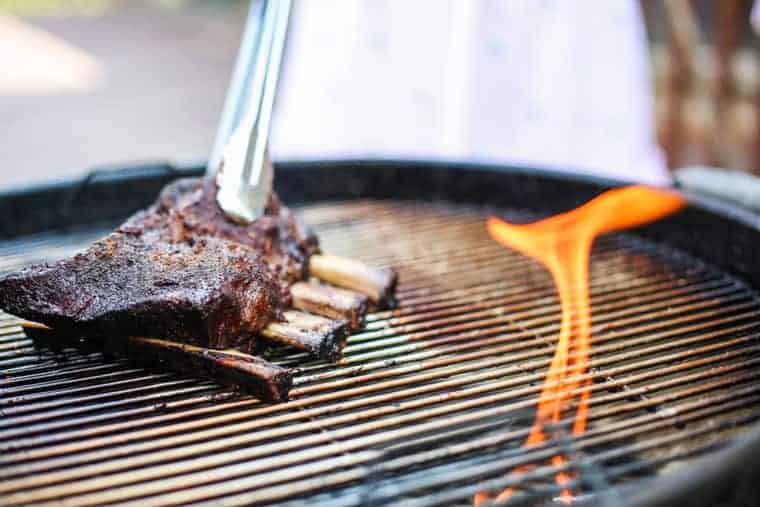 Beef ribs held with tongs over a BBQ grill next to a flame on the right coming up from burning charcoal