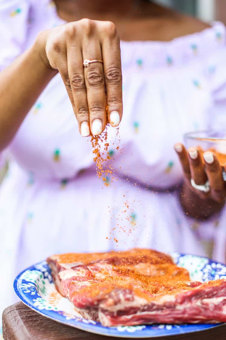 Raw beef ribs sitting on a blue and white plate being sprinkled with seasoning before grilling