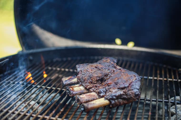 Charred beef ribs ready to eat is getting the finishing touches on a grill with flames coming up from the charcoal on the left