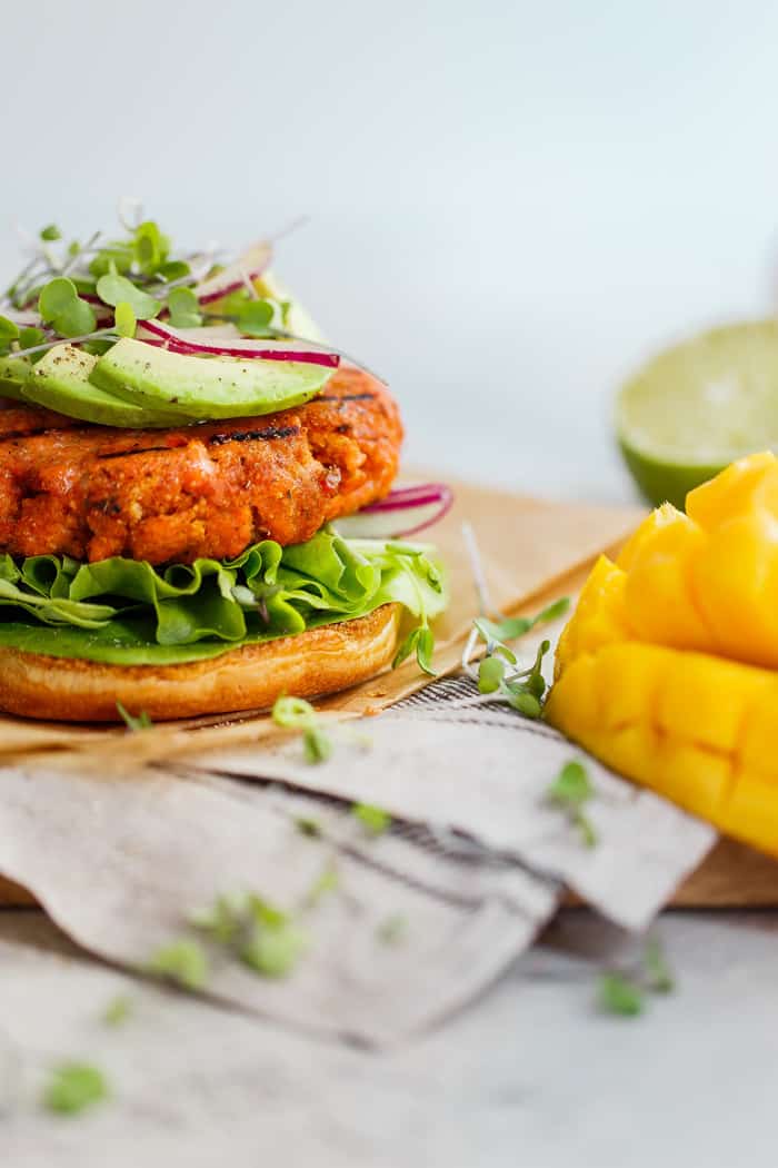 Mango jerk salmon burger served on a wooden cutting board next to a sliced mango and topped with lettuce, sprout and radish