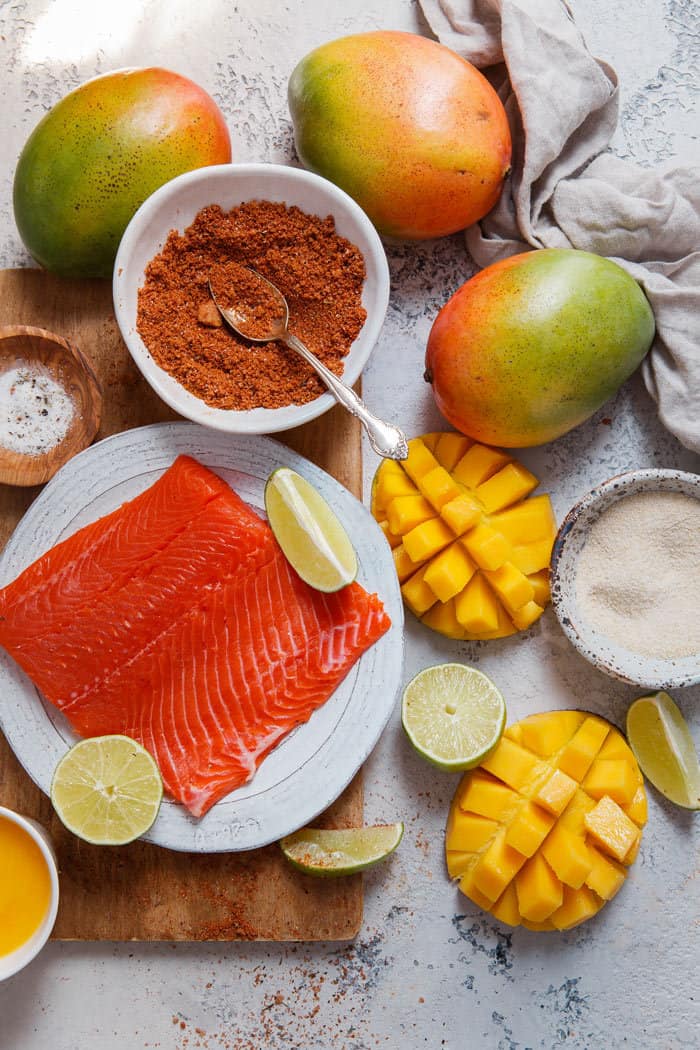 Overhead shot of all the ingredients needed to make this mango jerk salmon burger including jerk seasoning, raw salmon, sliced mangoes and limes all on a wooden cutting board 