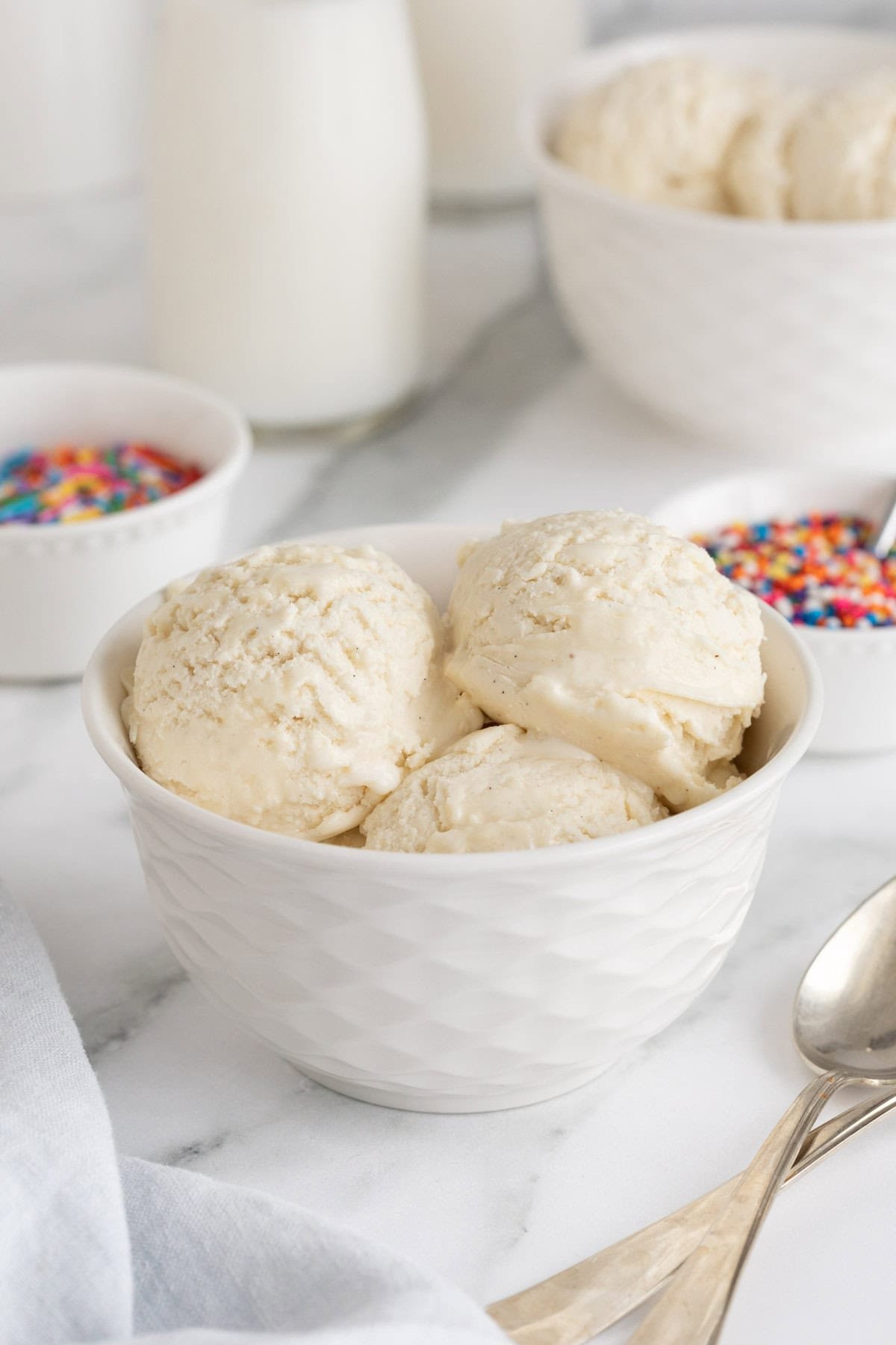Bowl of homemade vanilla ice cream with three scoops, surrounded by jars of sprinkles and bottles of milk