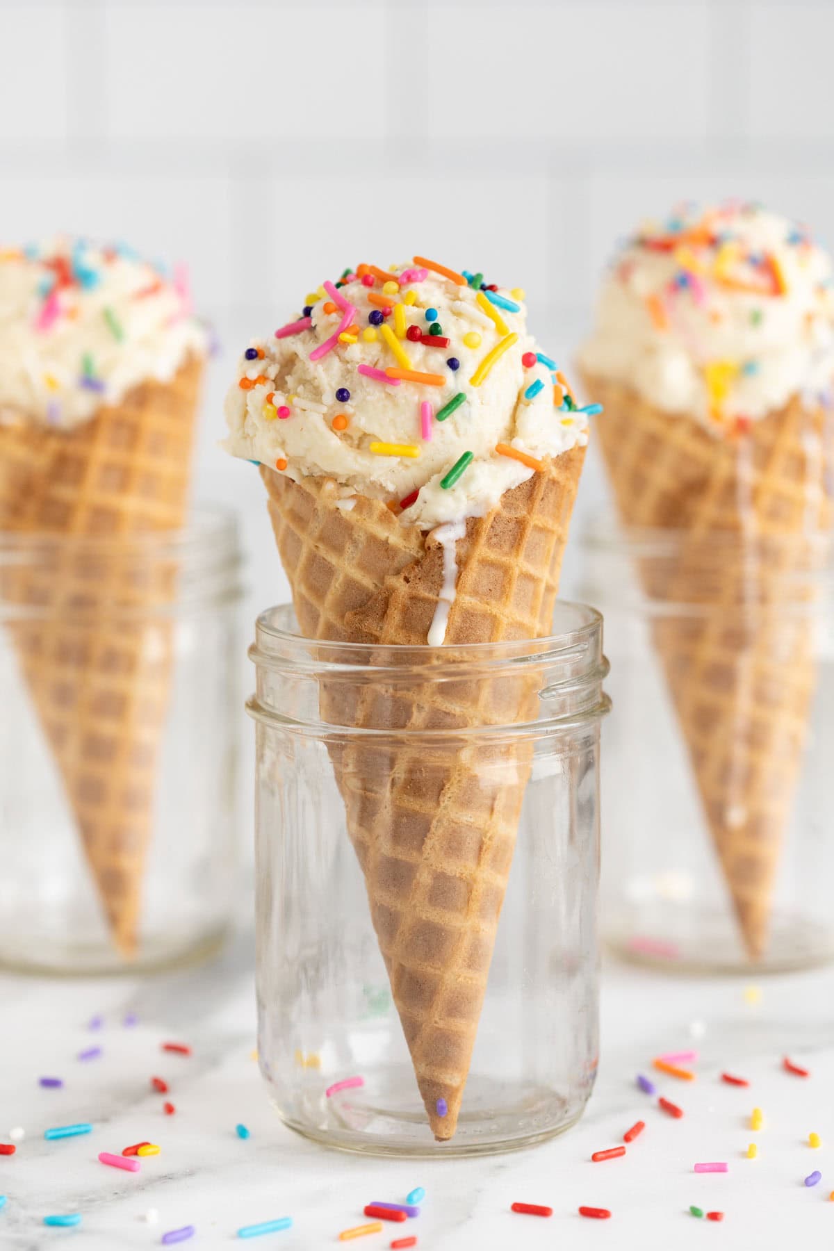 Waffle cone filled with homemade vanilla ice cream and rainbow sprinkles, standing upright in a glass jar with more cones in the background